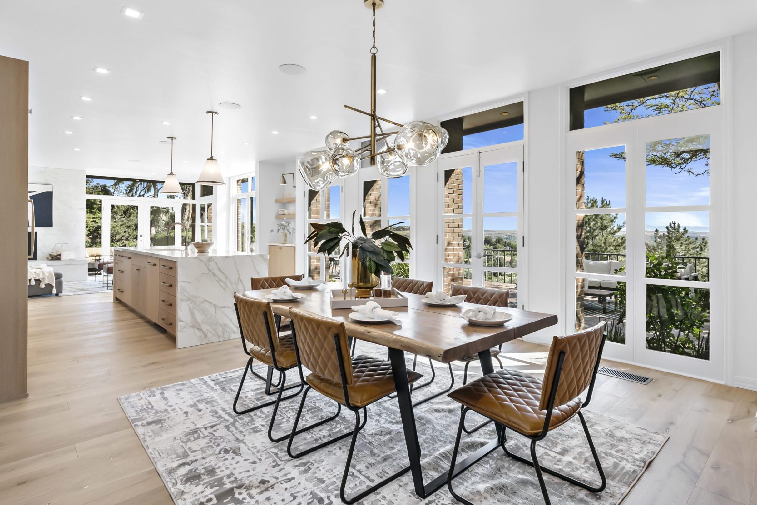 Stunning luxury interior view of dining space into the modern kitchen. Bright white wall of windows and french doors that lead to the back patio and overlook of the Treasure Valley